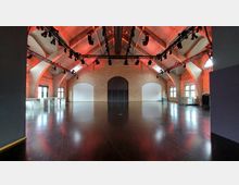 Großer Veranstaltungsraum mit hohen Gewölbedecken aus Beton, Backsteinwänden und dunkel glänzendem Holzboden. An der Decke ist ein modernes Beleuchtungssystem installiert, und der Raum verfügt über mehrere Fenster mit weißen Rahmen sowie eine zentrale große Doppeltür.