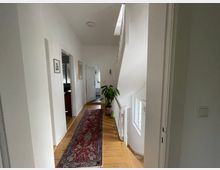 Ein heller Hausflur mit Holzfußboden und einem langen roten Läuferteppich, links führen Türen in angrenzende Räume, rechts sind weiße Treppengeländer mit einer Treppe, die nach unten führt. Im Hintergrund steht eine große grüne Zimmerpflanze vor einer weißen Wand.