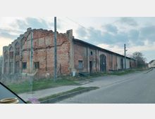 Altes, verlassenes Backsteingebäude mit beschädigten Wänden und teils zugemauerten Fenstern, neben einer gepflasterten Straße in einer ländlichen Umgebung. Im Hintergrund sind Strommasten und vereinzelte Häuser erkennbar.