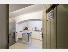 Blick in einen schlichten Hauswirtschaftsraum mit weißen Fliesen und gewölbter Decke. Im Raum befinden sich ein Kühlschrank, eine Spüle mit Unterschränken, ein Wäschetrocknergestell mit Handtüchern und verschiedene Haushaltsgegenstände.