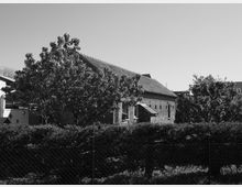 Schwarz-Weiß-Aufnahme eines einstöckigen Ziegelhauses mit Satteldach, umgeben von Bäumen und Hecken. Im Vordergrund verläuft ein Maschendrahtzaun.