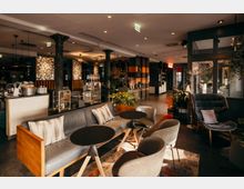 Moderner Lounge- und Cafébereich mit Sofas, gepolsterten Sesseln und runden Beistelltischen auf einem Teppich. Im Hintergrund stehen eine Bar-/Kaffeetheke mit Glasvitrinen, Regale, schwarze Säulen, Pflanzen und warme Deckenbeleuchtung in einem offenen Innenraum.