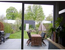 Blick durch eine große Glasschiebetür auf eine Terrasse mit grauen Fliesen, einem Holztisch mit Stühlen und einem geschlossenen Sonnenschirm. Dahinter liegt ein Garten mit grüner Rasenfläche, Büschen, blühenden Bäumen und einem modernen, flachen Gebäude mit weißer Tür im Hintergrund.
