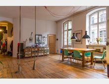 Offene Wohnküche in einem Altbau mit Holzboden, großen Fenstern und weißen Wänden. Im Raum befinden sich ein rustikaler Holztisch mit bunten Stühlen, eine Schaukel an der Decke, eine Kommode im Vintage-Stil sowie hängende Lampen und Dekorationsgegenstände.