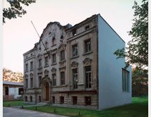 Historisches Gebäude mit verziertem Stuck an der Fassade auf der linken Seite und modernen, glatten Wänden auf der rechten Seite. Das Gebäude steht auf einem grünen Rasen in einer ruhigen, baumreichen Umgebung.