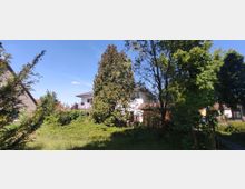 Ein weißes Wohnhaus mit Flachdach, teilweise verdeckt durch hohe Bäume und Sträucher in einem Garten. Im Vordergrund befindet sich eine Grünfläche, während links und rechts Nebengebäude und Zäune sichtbar sind.