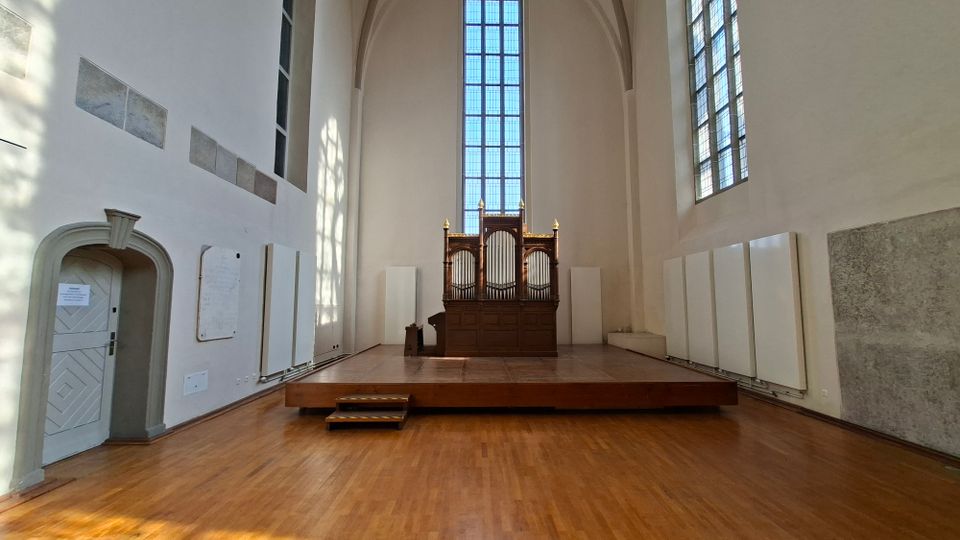 Heller Kirchen- oder Kapelleninnenraum mit hoher Gewölbedecke, großem hohen Fenster und einer Orgel auf einer erhöhten Holzplattform. Weiße Wände mit Akustikpaneelen und Holzboden prägen den Raum, seitlich sind weitere hohe Fenster und eine Tür sichtbar.