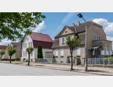 Straßenszene mit Einfamilienhäusern im traditionellen Stil, gekennzeichnet durch Satteldächer mit roten und braunen Ziegeln. Die Gebäude haben verputzte Fassaden, teils mit weißen Fensterläden, und sind von kleinen Vorgärten mit Bäumen und Zaunanlagen umgeben.