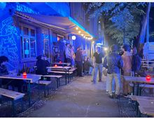 Außenbereich einer Bar an einer städtischen Straße bei Nacht, mit Holz-Bierzeltgarnituren und roten Kerzenlichtern unter einer Markise und Lichterkette. Die blau beleuchtete Fassade ist mit Graffiti versehen, daneben stehen Bäume und ein geparkter Transporter am Straßenrand.