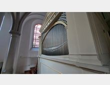 Nahaufnahme einer Kirchenorgel in einem weiß gestrichenen Gehäuse mit goldenen Details. Im Hintergrund ist ein großes Buntglasfenster und ein Teil der gewölbten Architektur der Kirche zu sehen.