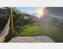 Garten eines Wohnhauses mit großer Rasenfläche, kleinen Bäumen und Hecken, im Vordergrund ein Kiesbett und ein Holzpfosten mit aufgehängter Hängematte. Rechts steht das Haus mit Terrasse und Hochbeet, im Hintergrund weitere Wohnhäuser im Abendlicht.