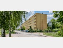 Wohngebäude mit gelber Fassade und mehreren Fenstern entlang einer ruhigen Straße, umgeben von Bäumen und parkenden Autos. Rechts im Bild steht ein Schild mit der Aufschrift „Gewerbepark Hopfenstr. 25“, umgeben von grünem Bewuchs.