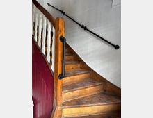 Holztreppe mit abgenutzten Stufen, einem hölzernen Geländer mit weißen Sprossen auf der linken Seite und einem modernen schwarzen Handlauf an der Wand rechts. Die Wand ist weiß, während der Bereich unter der Treppe in dunkelrot gestrichen ist.