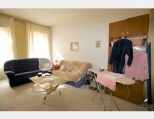 Wohnzimmer mit zwei Sofas, einem Glastisch mit goldfarbenem Gestell und heller Teppichboden. Rechts steht ein Bügelbrett mit Kleidung, daneben ein Holzschrank; die hellen Gardinen lassen Tageslicht herein.