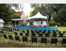 Bestuhlter Gartenbereich mit Reihen aus schwarzen und blauen Stühlen vor einer kleinen Holzbühne unter einem weißen Pavillonzelt, ausgestattet mit Mikrofonständern und Lautsprechern. Im Hintergrund stehen ein weißes Gartenhaus, ein Wohnhaus mit rotem Ziegeldach und hohe Bäume, die den Veranstaltungsplatz umgeben.