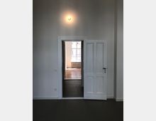 Innenansicht eines hellen Raums mit weißer Wand, einer geöffneten weißen Tür und Sicht auf einen angrenzenden Raum mit einem großen Fenster und einem Heizkörper. Das Licht über der Tür beleuchtet die Szene dezent.