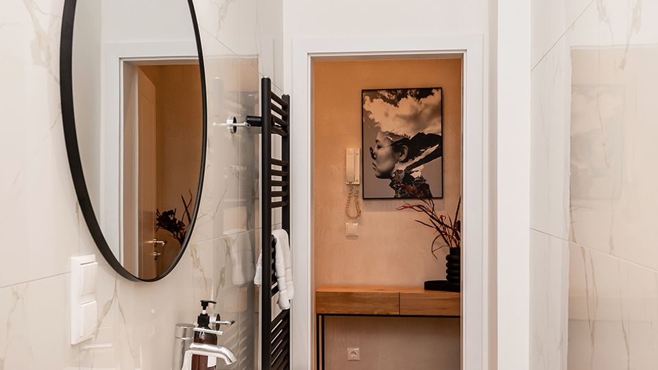 Schmales, modernes Badezimmer mit großformatigen weißen Marmorfliesen, grauem Boden und einem schwarzen Waschtischunterschrank mit rechteckigem Waschbecken und rundem Wandspiegel. An der Wand sind eine lineare LED-Leuchte und ein schwarzer Handtuchheizkörper zu sehen; am Ende öffnet sich der Raum zu einem Flur mit Holzkonsole und Wandbild.