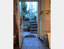 Blick durch eine geöffnete Tür in einen kleinen gepflasterten Hof mit Treppe und Geländer. Im Hintergrund befinden sich Pflanzen, Blumenkästen und Elemente einer mehrstöckigen Gebäudefassade.