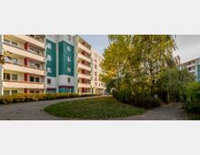 Mehrstöckiges Wohngebäude mit farbigen Fassadenabschnitten in Grün und Weiß, flankiert von einem Weg und Grünflächen mit Bäumen und Sträuchern im Vordergrund. Die Umgebung wirkt herbstlich, mit fallendem Laub und leichter Abendsonne.