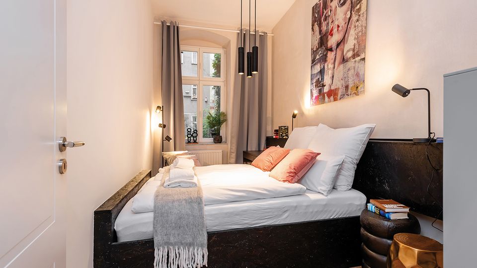 Schmales, modernes Schlafzimmer mit Doppelbett in dunklem Bettrahmen, weißer Bettwäsche und grauem Plaid sowie Nachttischen mit Leselampen. Am Ende des Raums steht ein großes Fenster mit grauen Vorhängen; darüber hängen schwarze Pendelleuchten und an der Wand ein großes Porträtbild.