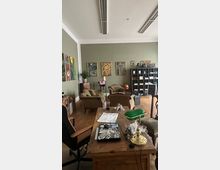 Ein Büro oder Arbeitsraum mit Holzboden, einem hölzernen Schreibtisch im Vordergrund und grüner Schreibtischlampe. Im Hintergrund befinden sich Sessel, ein Regal mit Fächern und dekorative Gemälde an einer olivgrünen Wand.