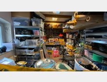 Ein kleiner Lagerraum mit Regalen voller Kunststoffboxen, Farbdosen, Werkzeugen und Bastelmaterialien. Im Raum gibt es eine Mischung aus Kartons, Arbeitsutensilien und Sitzgelegenheiten, sowie sichtbare Beleuchtung an der Decke.