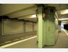 U-Bahnhof mit gefliesten Wänden, grauen Bodenfliesen und einem grünen, säulenartigen Konstrukt im Vordergrund. Im Hintergrund ist ein Schild mit der Aufschrift „Viktoria-Luise-Platz“ zu sehen, sowie ein Bahnsteig mit Sitzmöglichkeiten und Treppenaufgang.