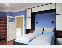 Schlafzimmer mit blau gestrichener Wand, einem Doppelbett mit blauer und weißer Bettwäsche und eingebauten weißen Schränken mit schwarzen Akzenten. Links steht eine geöffnete weiße Tür, daneben eine braun geflieste Heizwand und ein kleines Fenster.