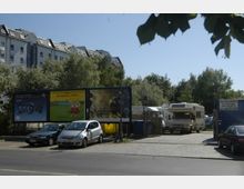 Straßenszene mit einem Parkplatz, auf dem mehrere Autos und ein Wohnmobil stehen. Im Hintergrund sind Werbetafeln, ein Zaun, sowie Bäume und ein mehrstöckiges Wohngebäude zu sehen.