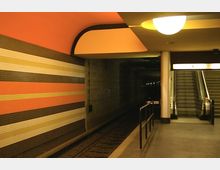 U-Bahn-Station mit einem Gleisbereich, einer farbigen Streifenmuster-Wand in Orange- und Beigetönen, einer Treppe und einer Rolltreppe auf der rechten Seite unter warmem Deckenlicht.