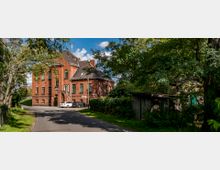 Backsteingebäude mit Satteldach und grünen Fensterrahmen, gelegen an einer ruhigen Straße mit parkenden Autos. Umgeben von Bäumen und Sträuchern, mit einem kleinen, hölzernen Schuppen auf der rechten Seite.