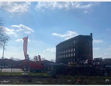Backstein-Gebäude mit mehreren Etagen neben einer Baustelle mit Baugeräten und gelagertem Material; im Vordergrund ein Zaun und ein Fahnenmast, im Hintergrund blauer Himmel mit lockeren Wolken.