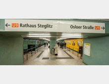 U-Bahn-Station mit grün gekachelten Wänden, einem Mittelgang und den Zugängen zu den Gleisen. Ein gelber U-Bahn-Zug der Linie U9 steht rechts, oben zeigen Schilder die Richtungen „Rathaus Steglitz“ und „Osloer Straße“ an.