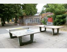 Zwei Tischtennisplatten aus Beton stehen auf einem gepflasterten Platz in einem Park. Im Hintergrund sind ein mit Graffiti bedecktes Backsteingebäude, Bäume und ein Spielplatz mit Schaukel und Rutsche zu sehen.