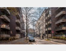 Straßenansicht mit mehrstöckigen Wohnhäusern aus Backstein, die Balkone mit Blumenkästen besitzen. In der Mitte der Straße stehen geparkte Autos, kahle Bäume und ein Straßenschild mit der Aufschrift „Charlottenburger Straße“.