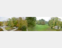 Weitläufiger Park mit grünem Rasen, umgeben von dicht bewachsenen Bäumen in herbstlichen Farben. Im rechten Bereich des Bildes ist ein kleines, helles Gebäude mit Säulen zu sehen, während links ein gepflasterter Weg und ein paar Personen erkennbar sind.