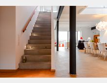 Moderne Wohnbereichskulisse mit einer geraden Betontreppe links, Hellholzböden und einem Essbereich mit einem großen Tisch und gepolsterten Stühlen rechts. Im Hintergrund sorgen große Fenster für viel Tageslicht, und mehrere dekorative Elemente, wie Lampen und eine auffällige Hängelampe, betonen das helle, offene Design des Raumes.