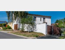 Zweistöckiges weiß verputztes Wohnhaus mit braunen Fensterrahmen und Türen, umgeben von einer weißen Mauer. Im Vordergrund stehen mehrere Bäume und Sträucher, im Hintergrund sind Nachbarhäuser und blauer Himmel zu sehen.