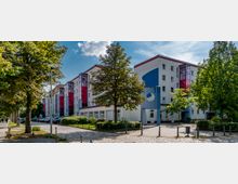 Mehrstöckiges Wohngebäude mit moderner, farbenfroher Fassade, geprägt von weißen Wänden, roten und blauen Details sowie gläsernen Treppenhäusern in Rot. Vor dem Gebäude befinden sich Bäume, Grünflächen, parkende Autos und ein gepflasterter Gehweg.