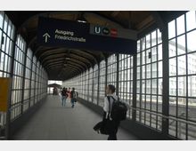 Ein überdachter Fußgängerübergang mit großen, vergitterten Fenstern auf beiden Seiten, der zu einem Ausgang mit der Beschilderung "Ausgang Friedrichstraße" führt. Mehrere Personen gehen in Richtung des Ausgangs, und rechts sind Schienen eines Bahnhofs zu sehen.
