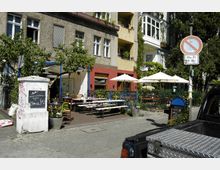 Straßenszene mit einem kleinen Café im Außenbereich vor einem mehrstöckigen Wohngebäude mit bunten Fassaden und Balkonen. Der Bereich ist mit Tischen, Bänken und Sonnenschirmen ausgestattet, umgeben von Pflanzen und einem Metallzaun.