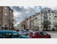 Straßenszene mit mehrstöckigen Wohngebäuden im Altbau- und Nachkriegsstil; helle Fassaden mit Balkonen und roten Dächern. Im Vordergrund parken mehrere Autos, und kahle Bäume stehen entlang der Gehwege unter einem bewölkten Himmel.