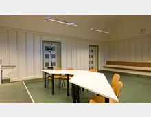 Ein schlichter, rechteckiger Besprechungsraum mit hellen Wänden und grünen Fußböden. Zentral steht ein weißer, unregelmäßig geformter Tisch mit Holzstühlen, an der Wand befinden sich eine Garderobe, eine Sitzbank und eine Uhr.