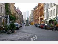 Straßenszene in einer Stadt mit historischen Gebäuden auf beiden Seiten, darunter ein Backsteinbau rechts. Gehwege sind mit Fahrrädern, geparkten Autos und Pflanzen dekoriert. In der Mitte steht ein Schild mit einem S-Bahn-Logo, im Hintergrund sind moderne Hochhäuser sichtbar.
