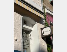 Fassade eines Gebäudes mit hellem Putz, einem gelb-weiß gestreiften Vordach, einem kleinen Fenster mit Gitter und einem alten Schild mit der Aufschrift „Videothek“ neben einer Illustration. Rechts im Bild wächst grüne Efeu-Pflanze an der Wand.