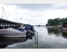 Ein Hafen an einem See mit mehreren festgemachten Booten, von denen einige mit Planen abgedeckt sind. Im Hintergrund sind weitere Boote sowie eine bewaldete Uferlandschaft zu sehen, während ein Steg und eine überdachte Anlegestelle den Vordergrund dominieren.