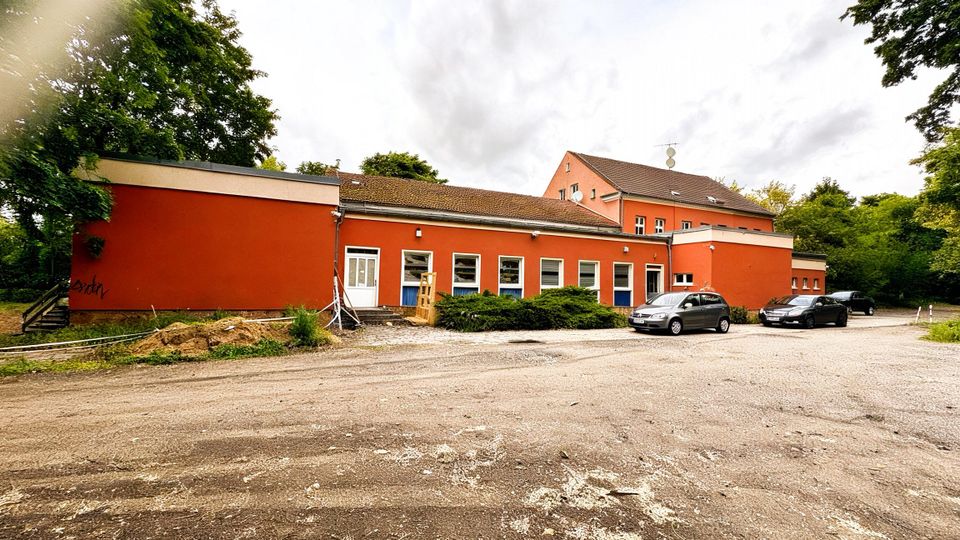 Flaches, langgezogenes rotes Gebäude mit vielen rechteckigen Fenstern und mehreren Eingängen, dahinter ein höherer Gebäudeteil mit Satteldach. Davor liegt ein großer, unbefestigter Parkplatz mit einigen abgestellten Autos, umgeben von Bäumen und Grünflächen.