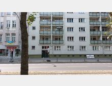 Mehrstöckiges Wohngebäude mit hellgrauer Fassade und Balkonen mit grünem Glasgeländer; im Erdgeschoss befindet sich eine Eingangstür. Links daneben ein weiteres Gebäude mit einer Geschäftsfassade und einer ukrainischen Flagge im Fenster. Im Vordergrund verlaufen eine Straße und ein Gehweg, teils durch Bäume verdeckt.