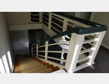 Treppenhaus mit hölzernen Stufen und einem cremefarbenen Geländer, das mit einem dunkelgrünen Handlauf versehen ist. Das Geländer hat horizontale Streben, und das Treppenhaus erhält teilweise Tageslicht durch eine nahegelegene Fensteröffnung.