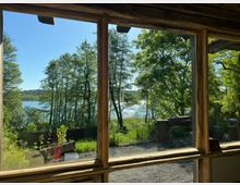 Blick durch ein Fenster mit hölzernem Rahmen auf eine Waldlandschaft mit Bäumen, einem See im Hintergrund und hellem Himmel. Im Vordergrund sind eine Bank und einige Gartenmöbel sichtbar, umgeben von Gras und Pflanzen.